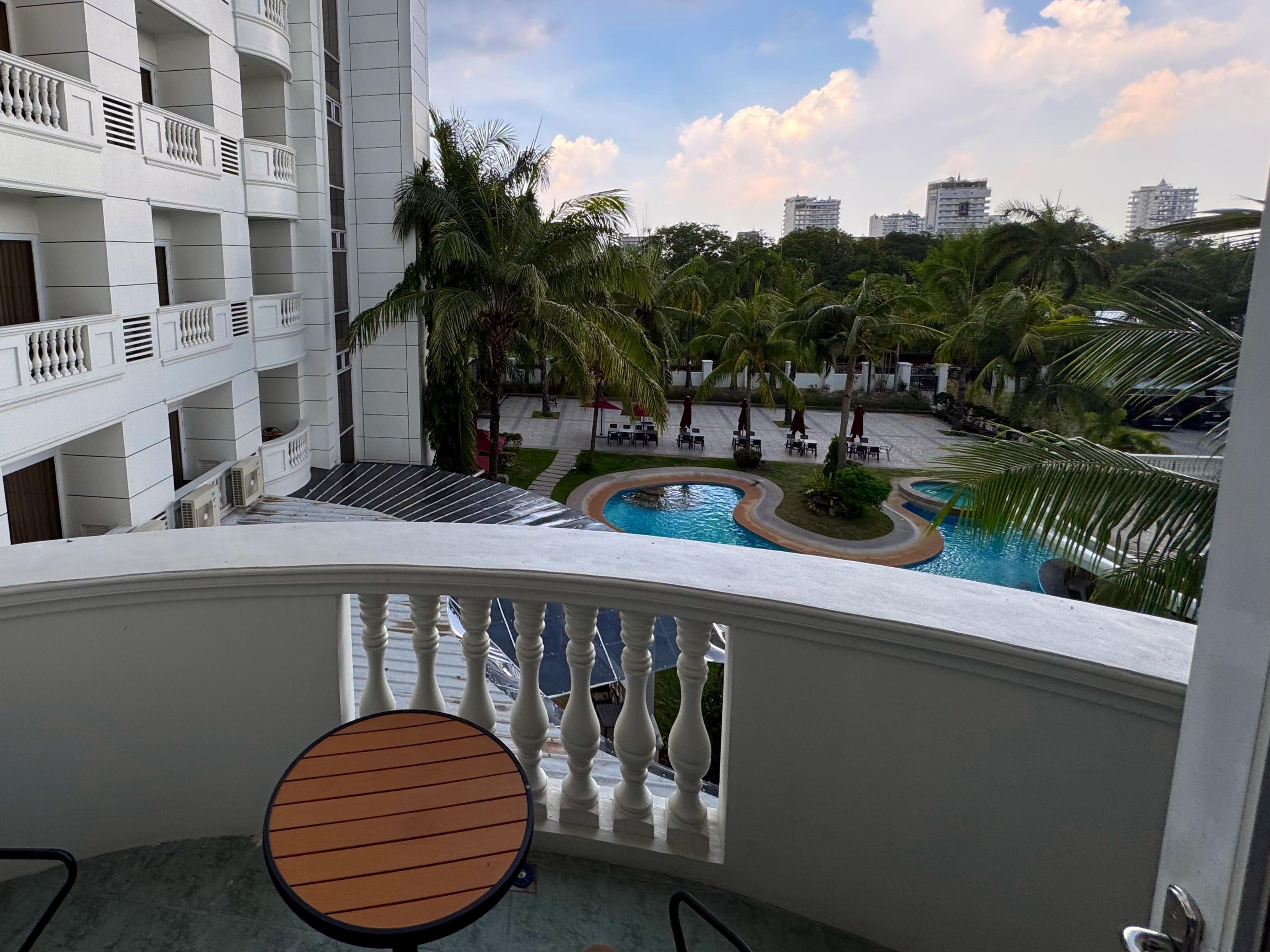 A balcony with a view of a swimming pool, palm trees, and a multi-story building.
