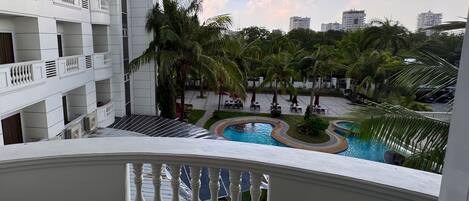 A balcony with a view of a swimming pool, palm trees, and a multi-story building.