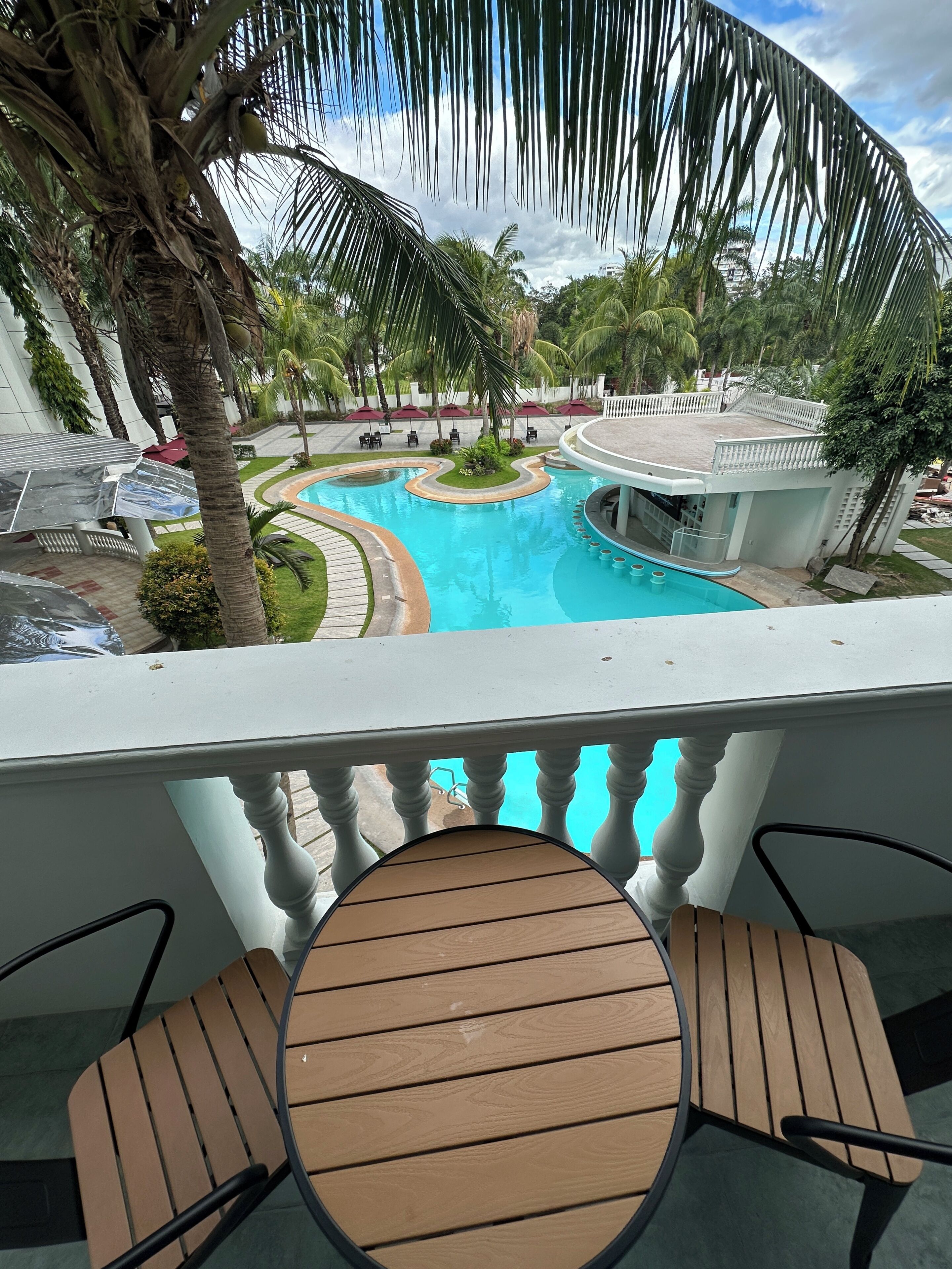 A pool with a curved design, surrounded by palm trees and a white building with a balcony.