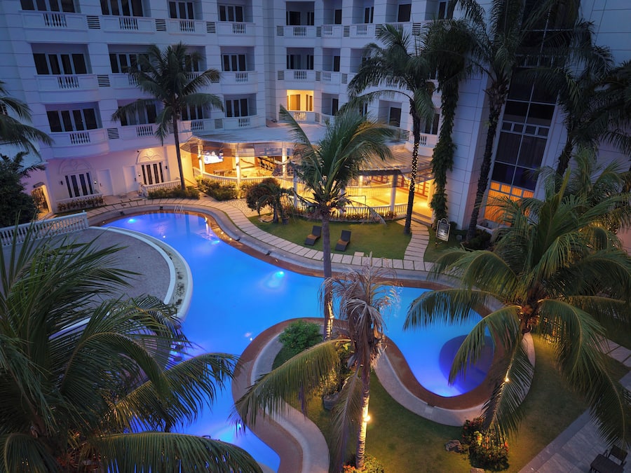 A swimming pool with a curved design, surrounded by palm trees and a building with balconies.