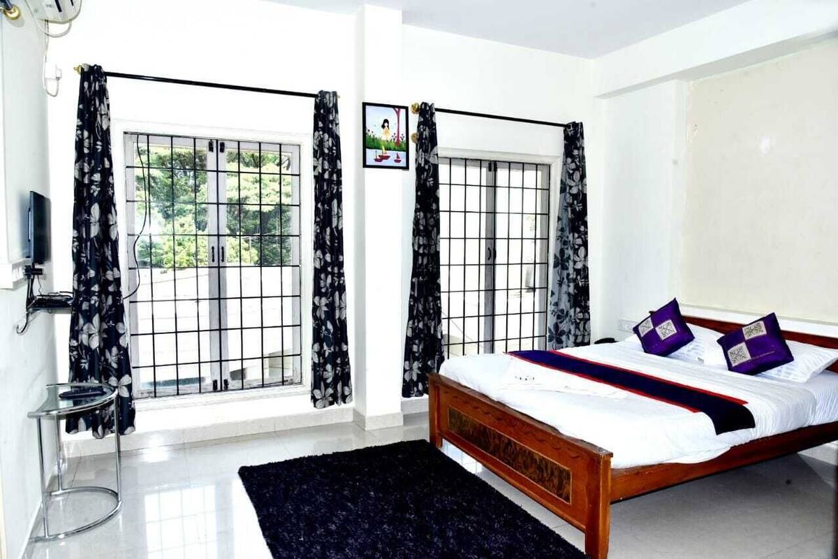 A bedroom with a bed, a TV, a small table, and two windows with curtains.