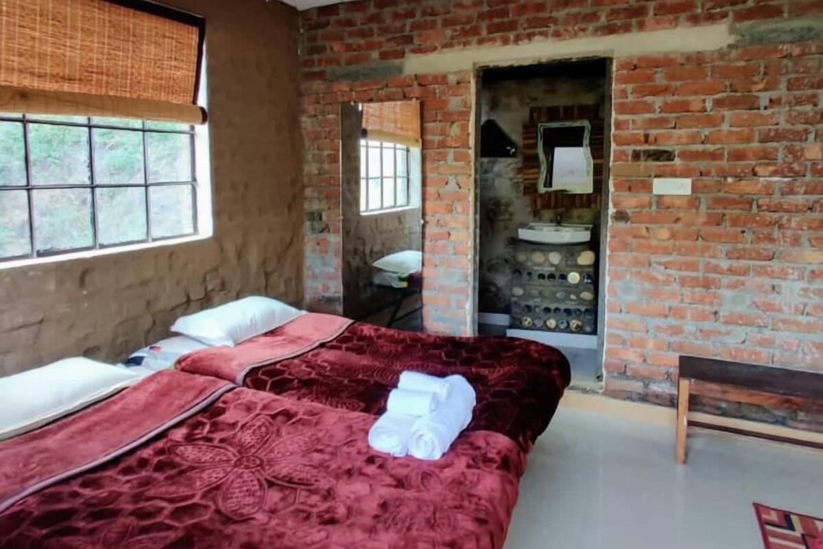 A room with a brick wall, a bed with red bedding, a window with a bamboo blind, and a small wooden bench.