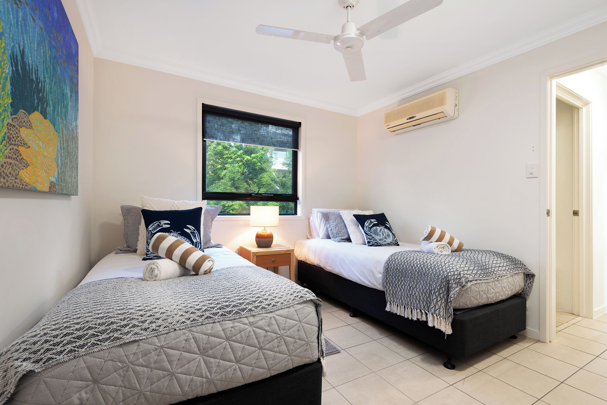 A bedroom with two beds, a ceiling fan, a window with a view of greenery, and an air conditioning unit.