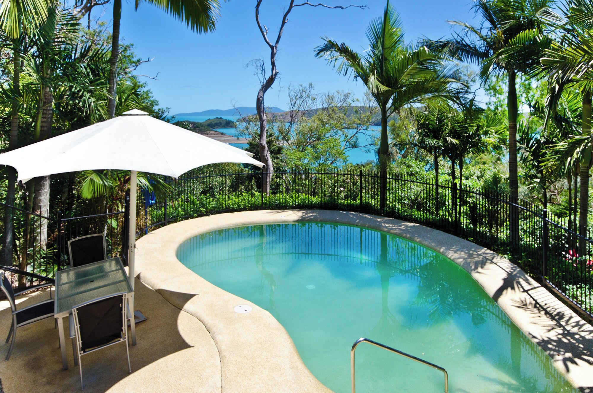 A curved pool with a white umbrella and metal chairs, surrounded by a black fence and lush greenery.