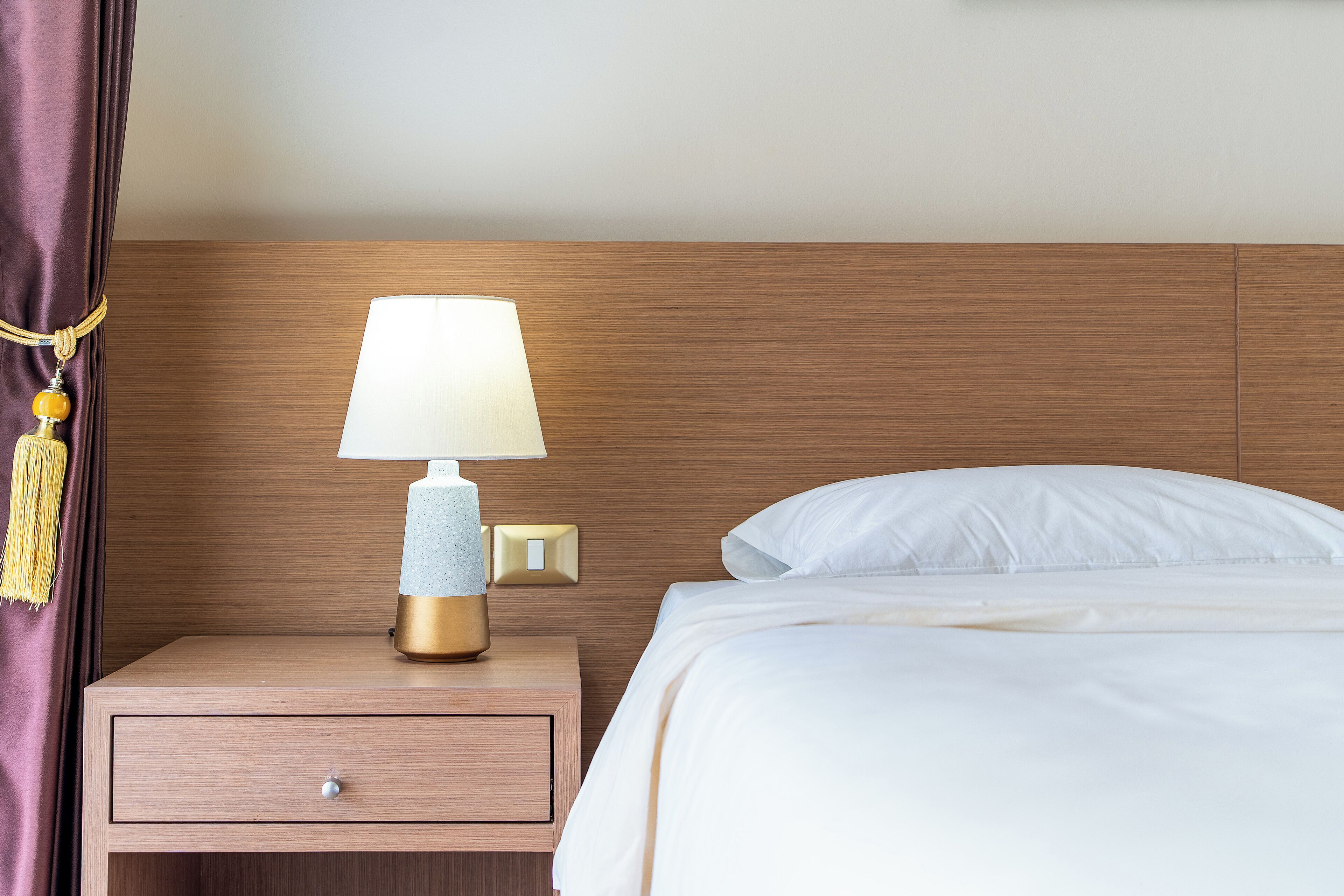 A neatly made bed with white linens, a wooden nightstand with a lamp, and a wall-mounted light switch.