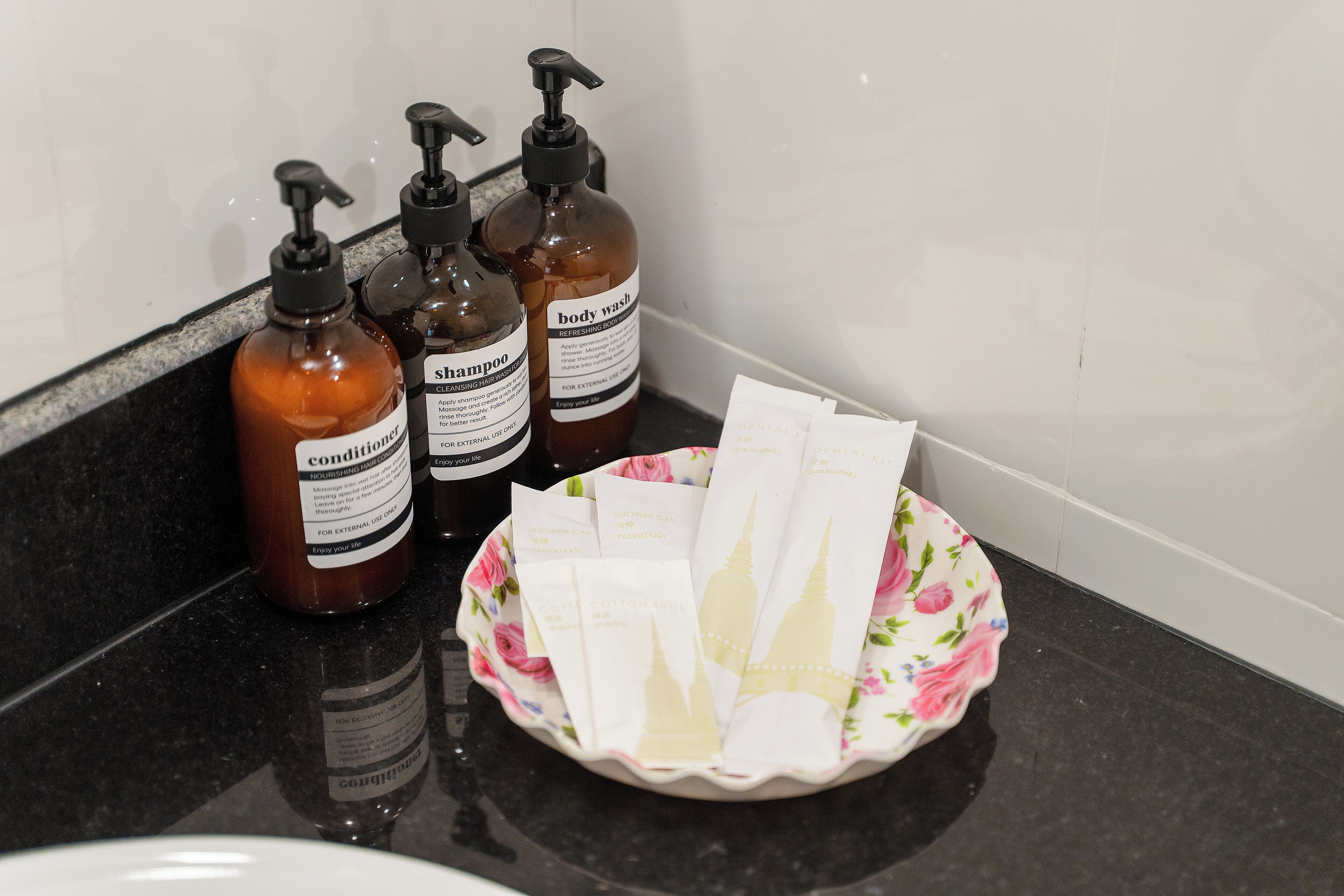 A bathroom counter with three bottles of shampoo and conditioner, and a bowl containing various toiletries.
