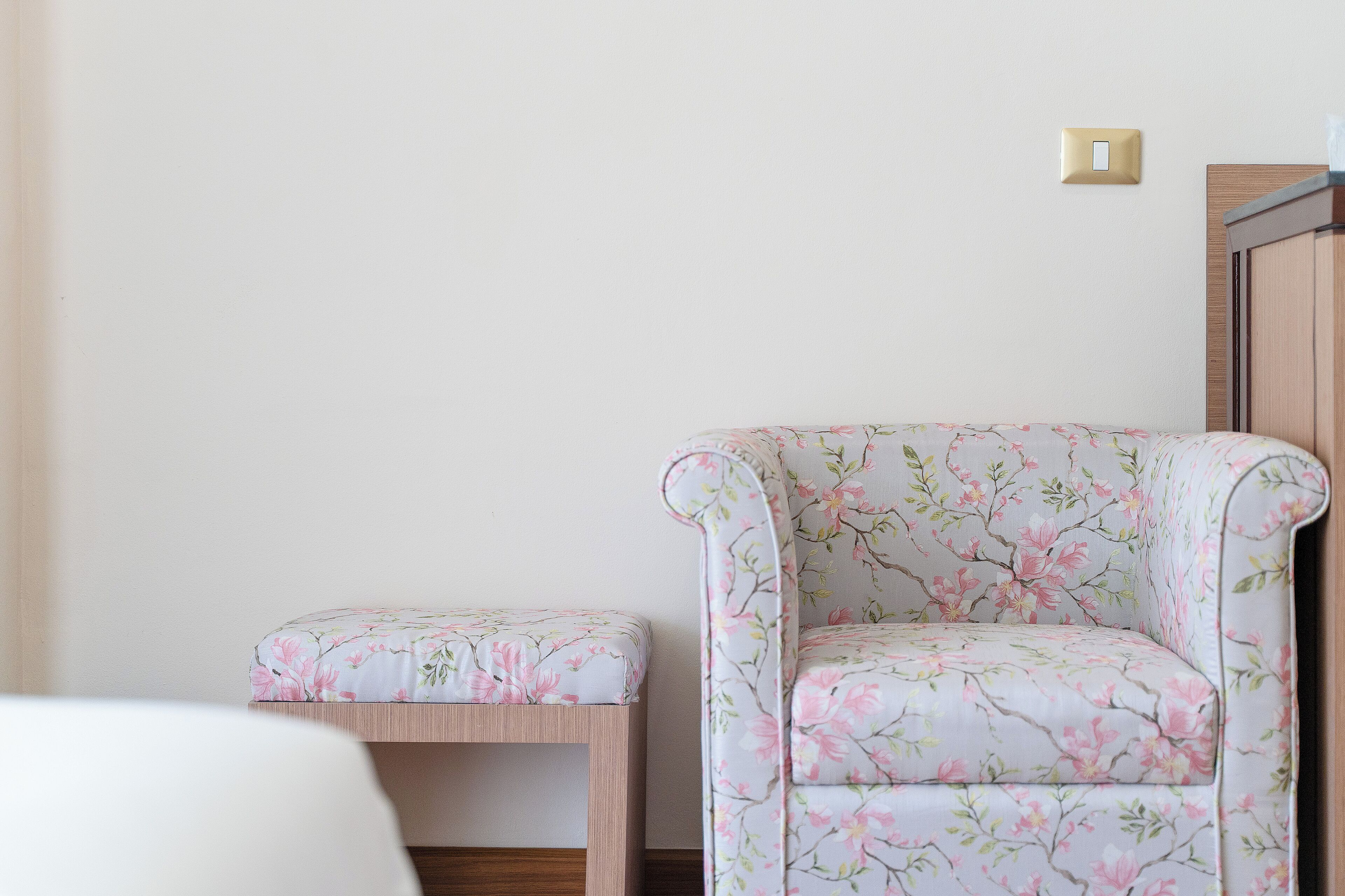 A room with a floral-patterned armchair and ottoman, a wooden side table, and a light switch on the wall.