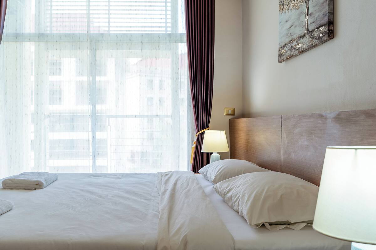 Een hotelkamer met een bed, een lamp, een raam met gordijnen en een schilderij aan de muur.