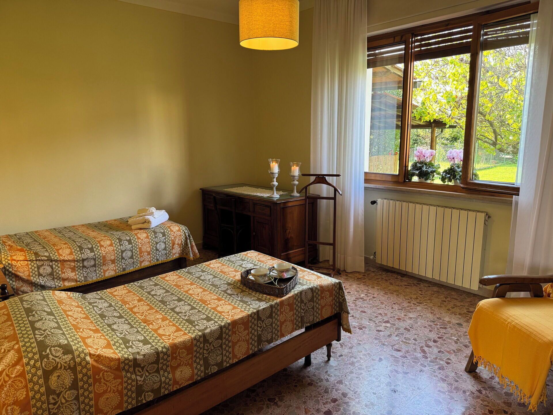 Une chambre avec deux lits, une commode en bois, un fauteuil suspendu, une fenêtre donnant sur la verdure et un radiateur.