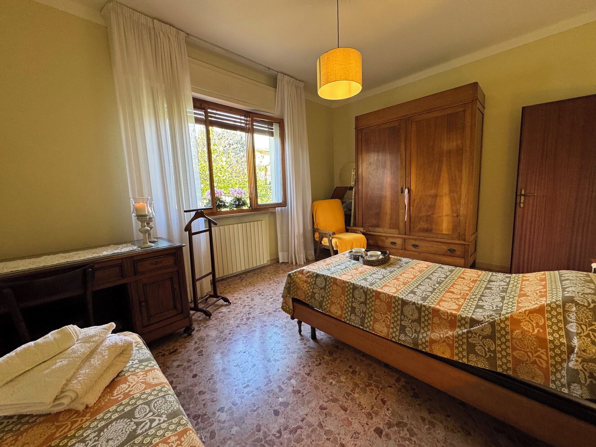 A bedroom with a bed, a wooden wardrobe, a desk, a chair, and a window with curtains.