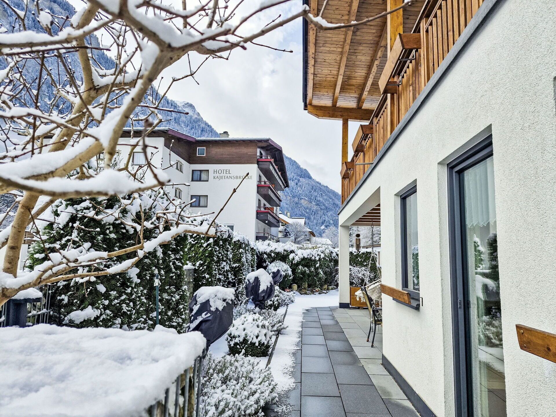 Ein mit Schnee bedeckter Hotelinnenhof mit einem Holzgebäude, einem Steinweg und Bergelandschaft im Hintergrund.