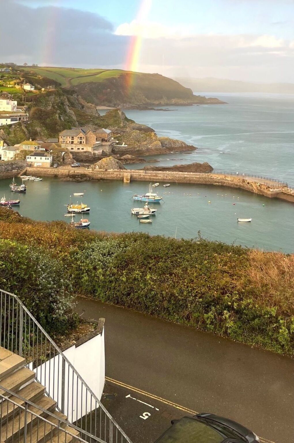 A coastal town with boats docked in a harbor, a rainbow in the sky, and buildings on the hillside.