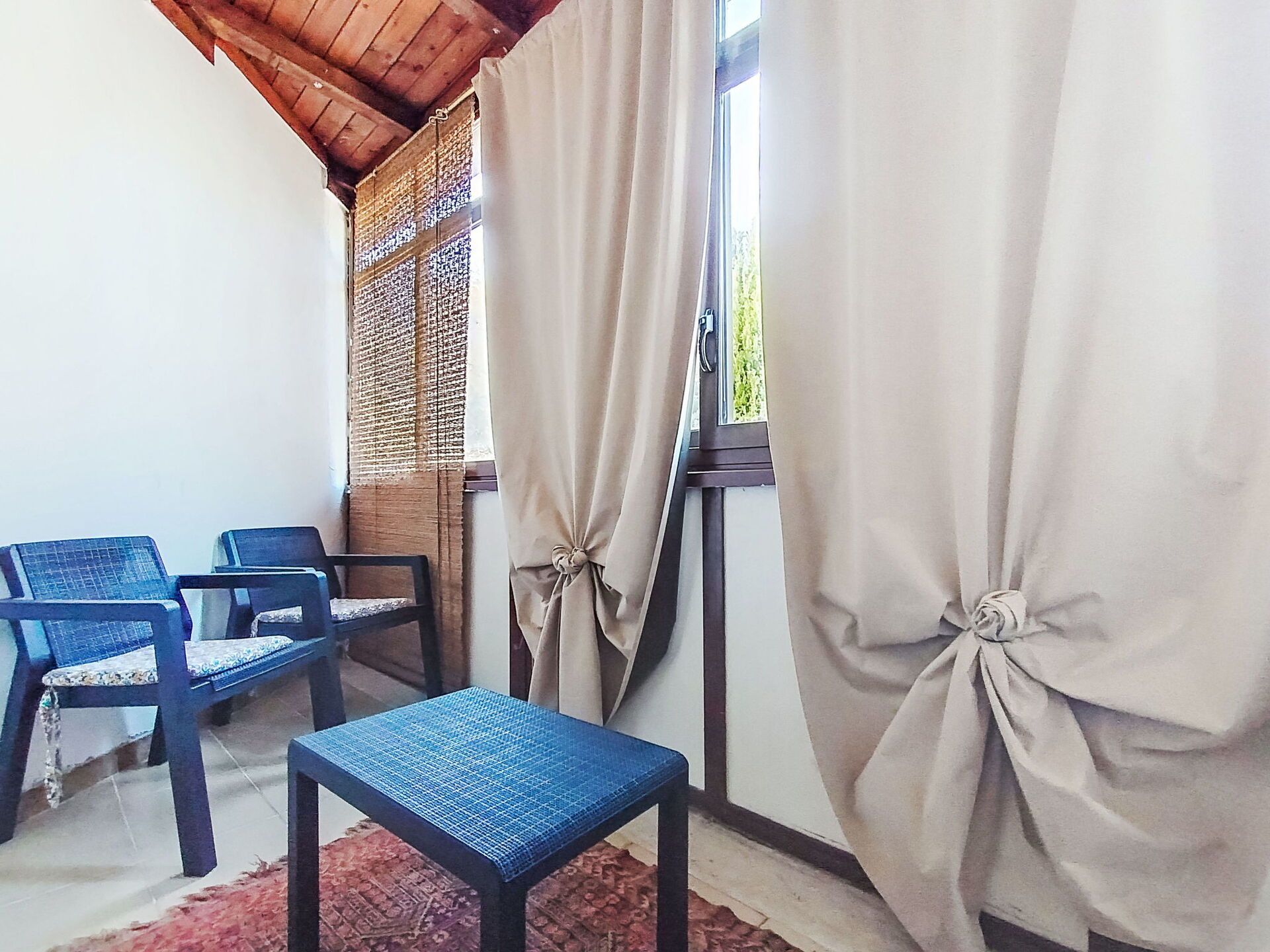 A room with two blue chairs, a small table, a window with curtains, and a wooden ceiling.