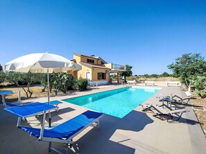 Ein Schwimmbecken mit Liegestühlen und Sonnenschirmen, ein gelbes Gebäude und ein klarer, blauer Himmel.