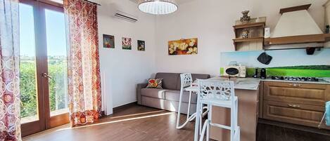 A modern kitchen with a dining area, a sofa, and a view of greenery outside.