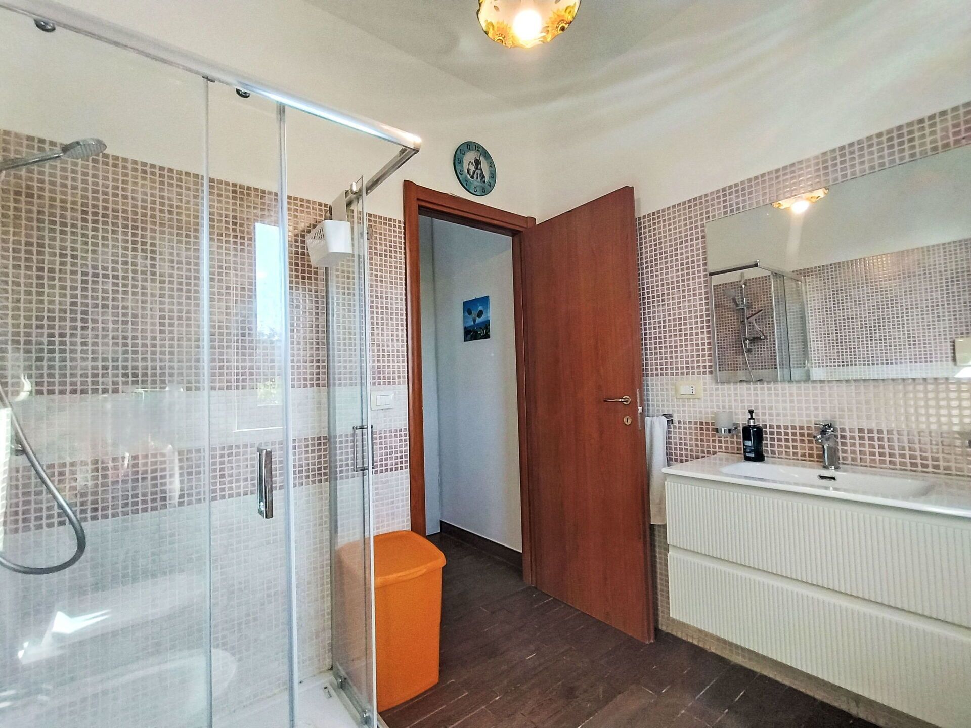 Ein Badezimmer mit einer gläsernen Duschkabine, einer Holztür, einem weißen Waschbecken inklusive Spiegel und einer gefliesten Wand.
