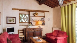 Una sala de estar acogedora con un sofá rojo, una mesa de centro de madera y un televisor. Se observa una estantería con libros y una lámpara. Se ve un ventanal con un diseño de celosía.