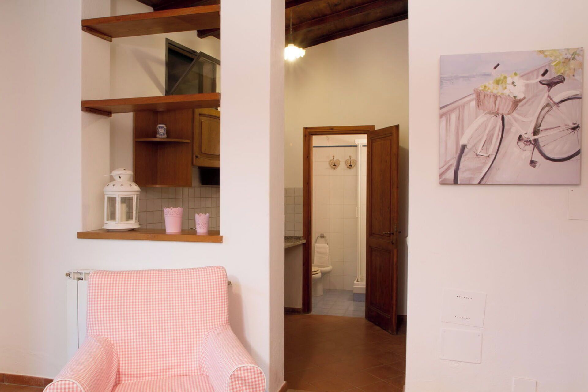 A room with a checkered chair, wooden shelves, a painting of a bicycle, and a view into a bathroom.