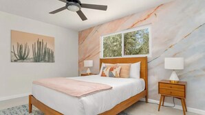 A bedroom with a bed, ceiling fan, marble wall, window, and two bedside tables with lamps.