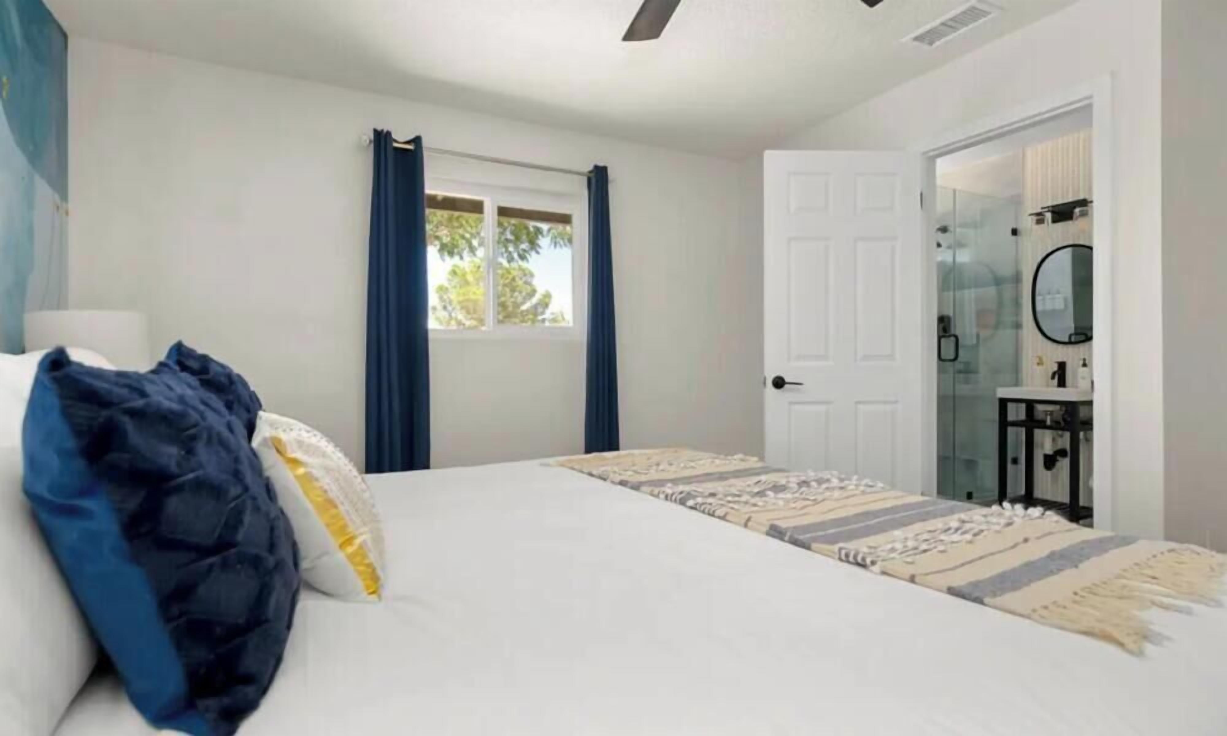 A bedroom with a bed, a window with curtains, a door, and a bathroom visible through a glass partition.