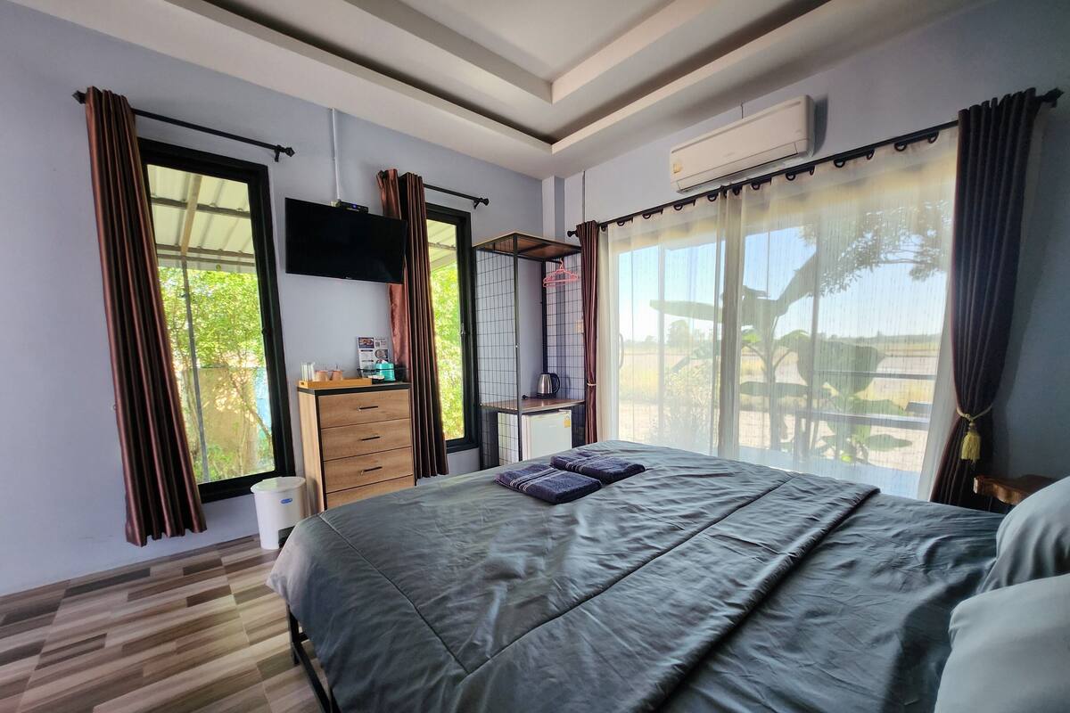 A bedroom with a large bed, a wooden dresser, a TV mounted on the wall, and a window with curtains.