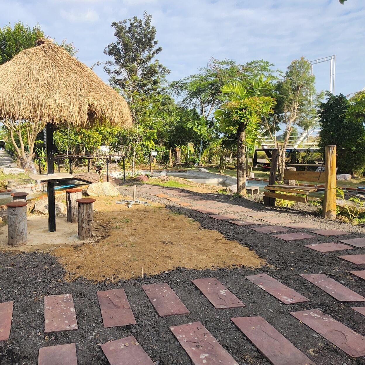 A thatched-roof structure with a bench and a small pool area, surrounded by trees and a paved walkway.