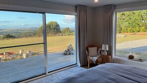 A bedroom with a large window offering a view of a grassy field and trees. There is a bed with a grey blanket, a wicker chair with a lamp, and a small table with a vase.