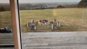 A view from a window overlooking a grassy field with a fire pit and logs, and a distant landscape.