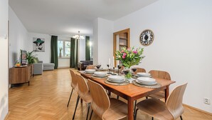 A dining area with a wooden table set for six, surrounded by modern chairs. A vase with flowers is on the table, and a clock is on the wall. - Lumina apartment for family in Warsaw (Warsaw)