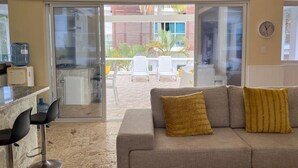 Ein Wohnzimmer mit einer Couch, zwei Sesseln, einem Wasserspender und einer Glastür, die auf eine Terrasse mit Tisch und Stühlen führt.
