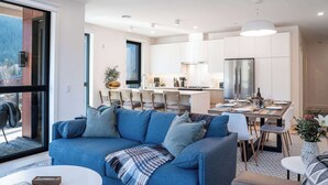 Een moderne woonkamer met een blauwe bank, een eethoek met een houten tafel en een keuken met witte kasten en een roestvrijstalen koelkast.