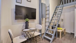 En moderne stue med et fladskærms-tv på væggen, et spisebord med stole, en lille potteplante og en trappe med gelænder i metal.