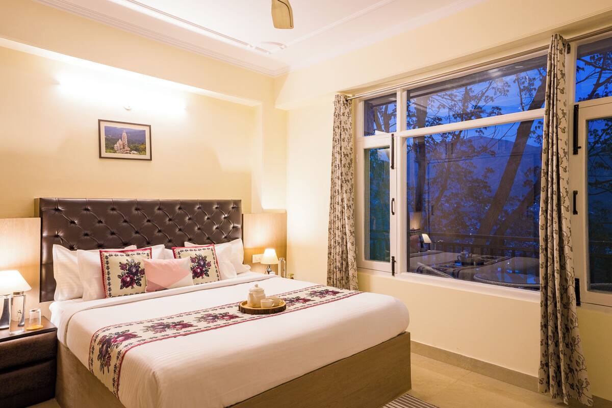 A well-lit bedroom with a large bed, a nightstand with a lamp, a window with curtains, and a framed picture on the wall.