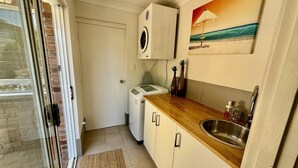 A compact kitchen with a built-in washing machine, a wooden countertop, a stainless steel sink, and a beach-themed painting on the wall.