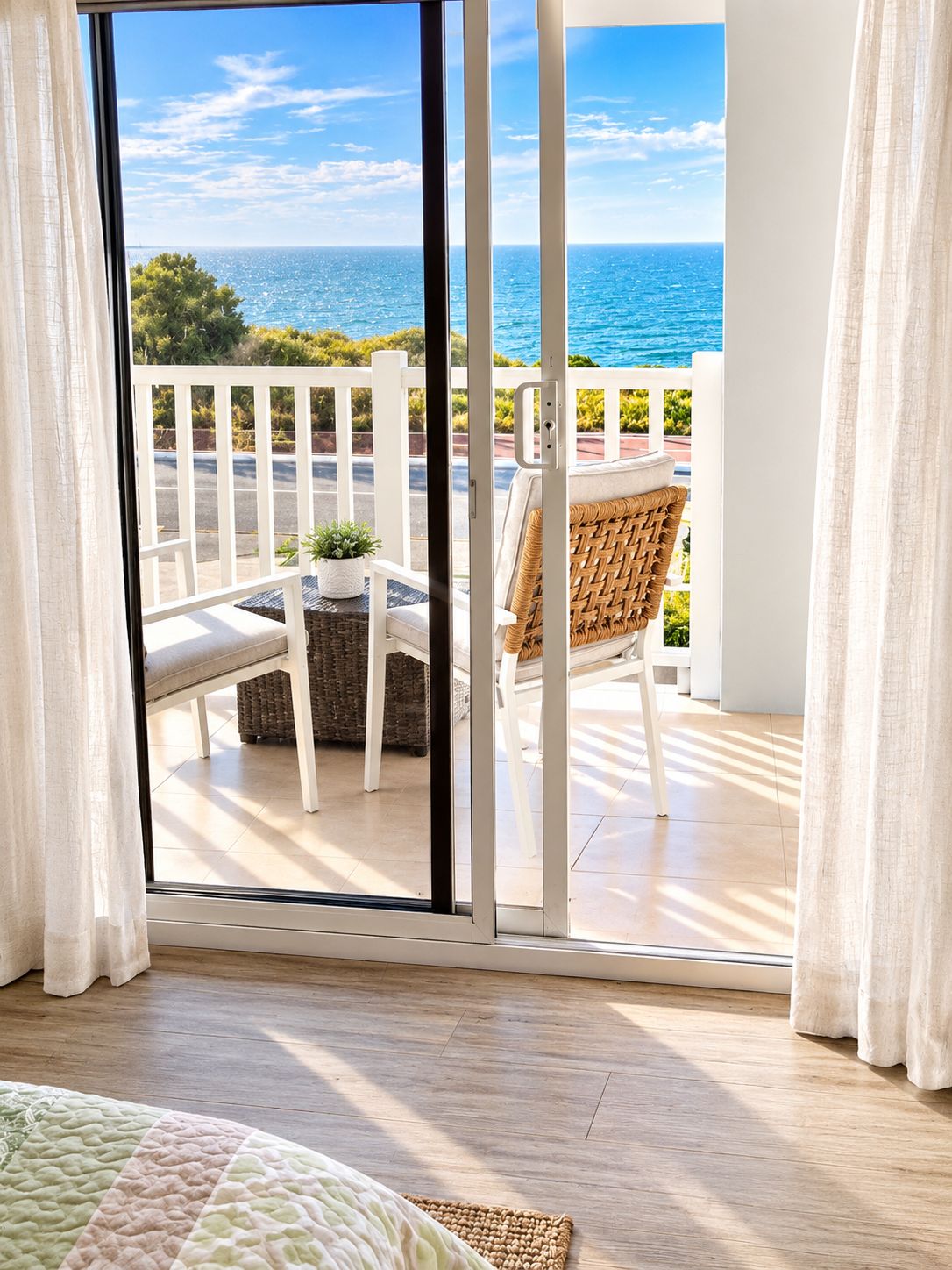 A room with a view of the sea through a sliding glass door, a wicker chair, a small table, and a white railing.