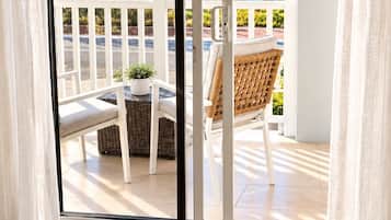 A room with a view of the sea through a sliding glass door, a wicker chair, a small table, and a white railing.