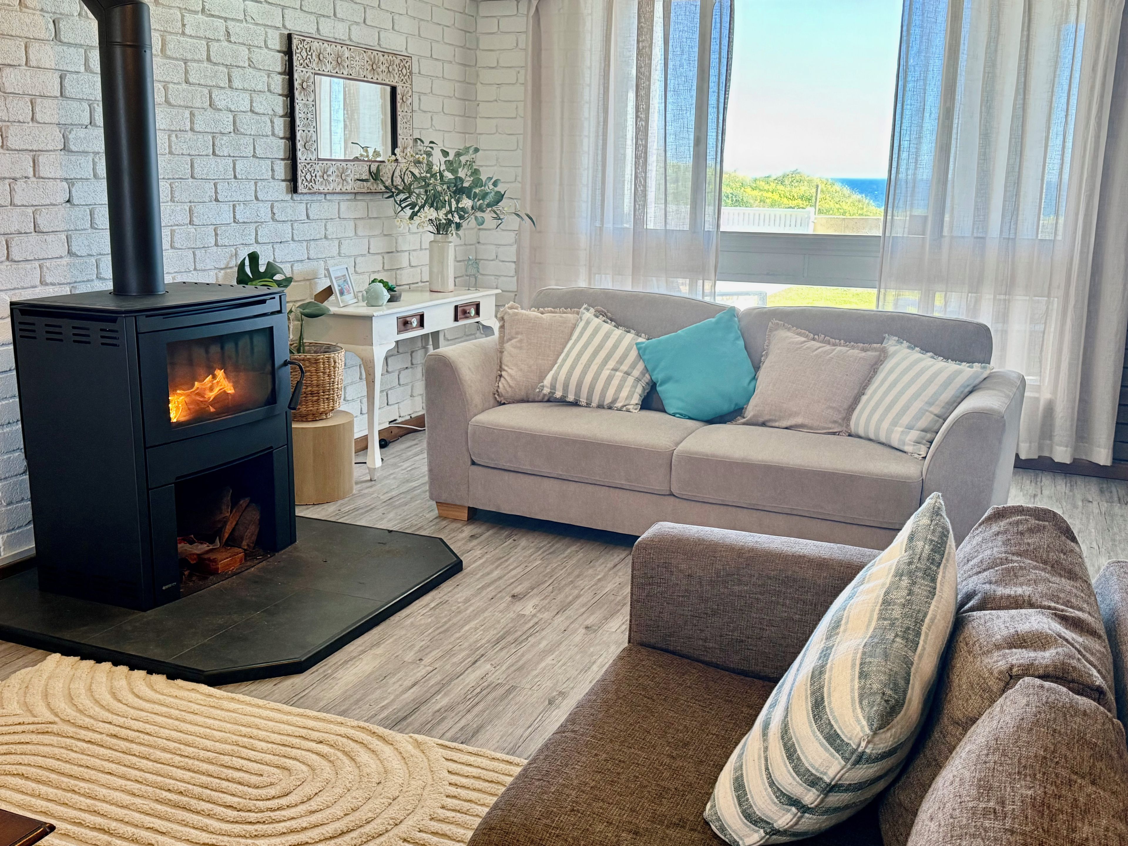 A living room with a black wood-burning stove, a beige sofa with pillows, a white desk with a plant, and a view through sheer curtains.