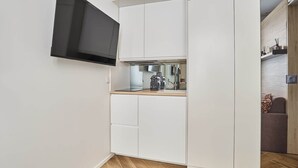 Cocina compacta con muebles blancos, microondas integrado y televisor de pantalla plana montado en la pared.