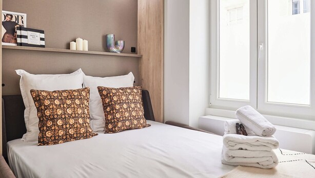 Una habitación de hotel con una cama, almohadas, una mesita de noche con una toalla, un estante con objetos decorativos y una ventana con vistas.