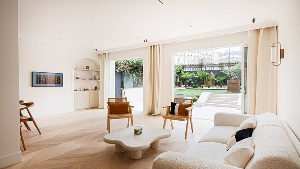 Een ruime woonkamer met een visgraatvloer, een beige bank, twee houten stoelen, een salontafel en uitzicht op een tuin door grote ramen.