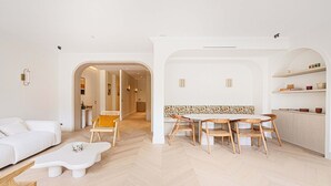 Een moderne woonkamer met een witte bank, een bijzondere witte salontafel, en een eethoek met een beklede bank en houten stoelen.