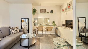 Un salon moderne avec un canapé, deux tables en verre et un coin cuisine équipé de tabourets de bar.