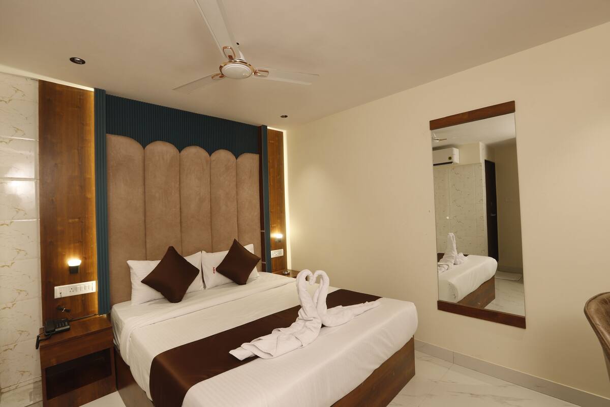 Habitación de hotel con una cama grande, un ventilador de techo, una mesita de noche, un espejo y un baño que se ve en el reflejo.