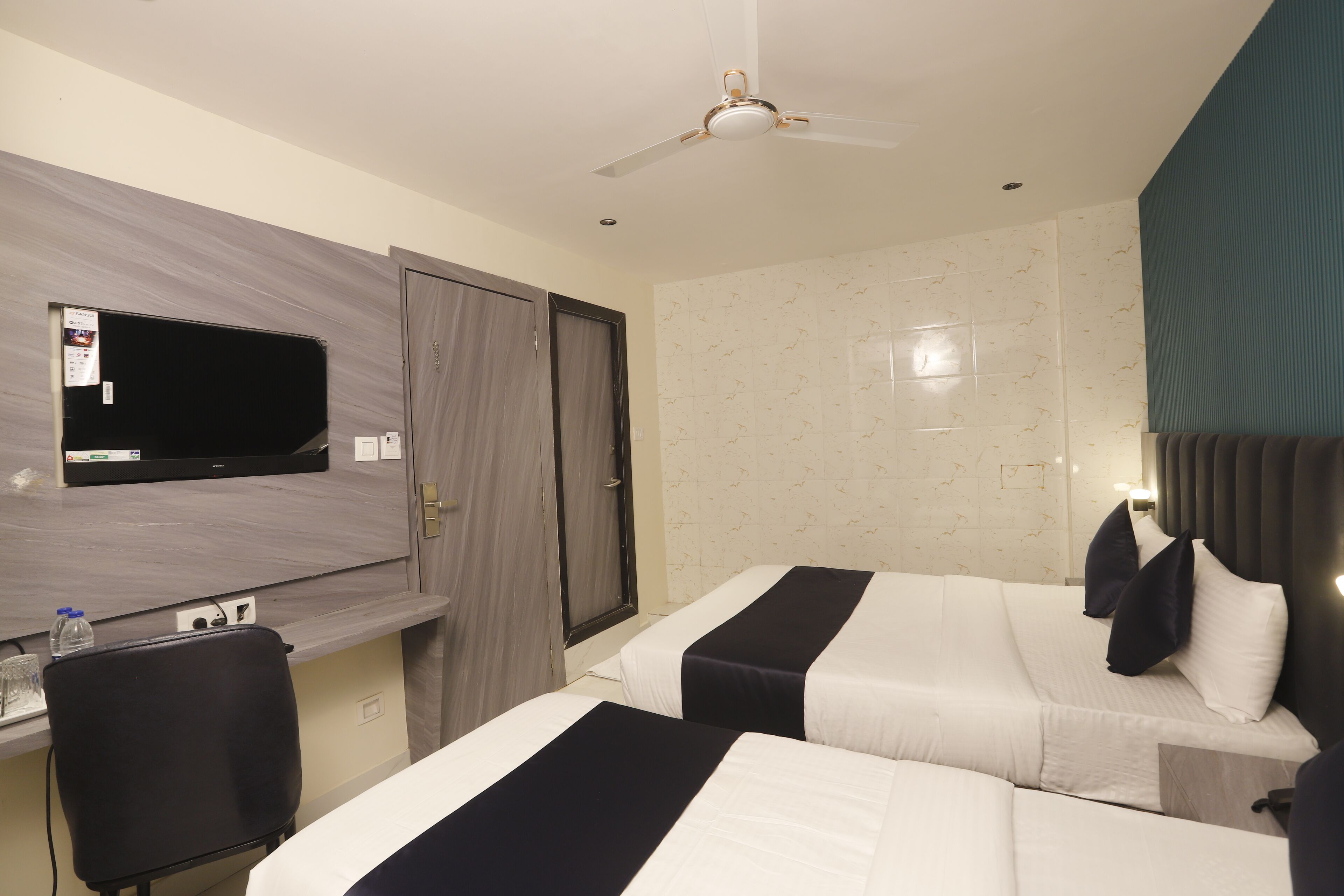 A hotel room with two beds, a desk, a television, and a ceiling fan.