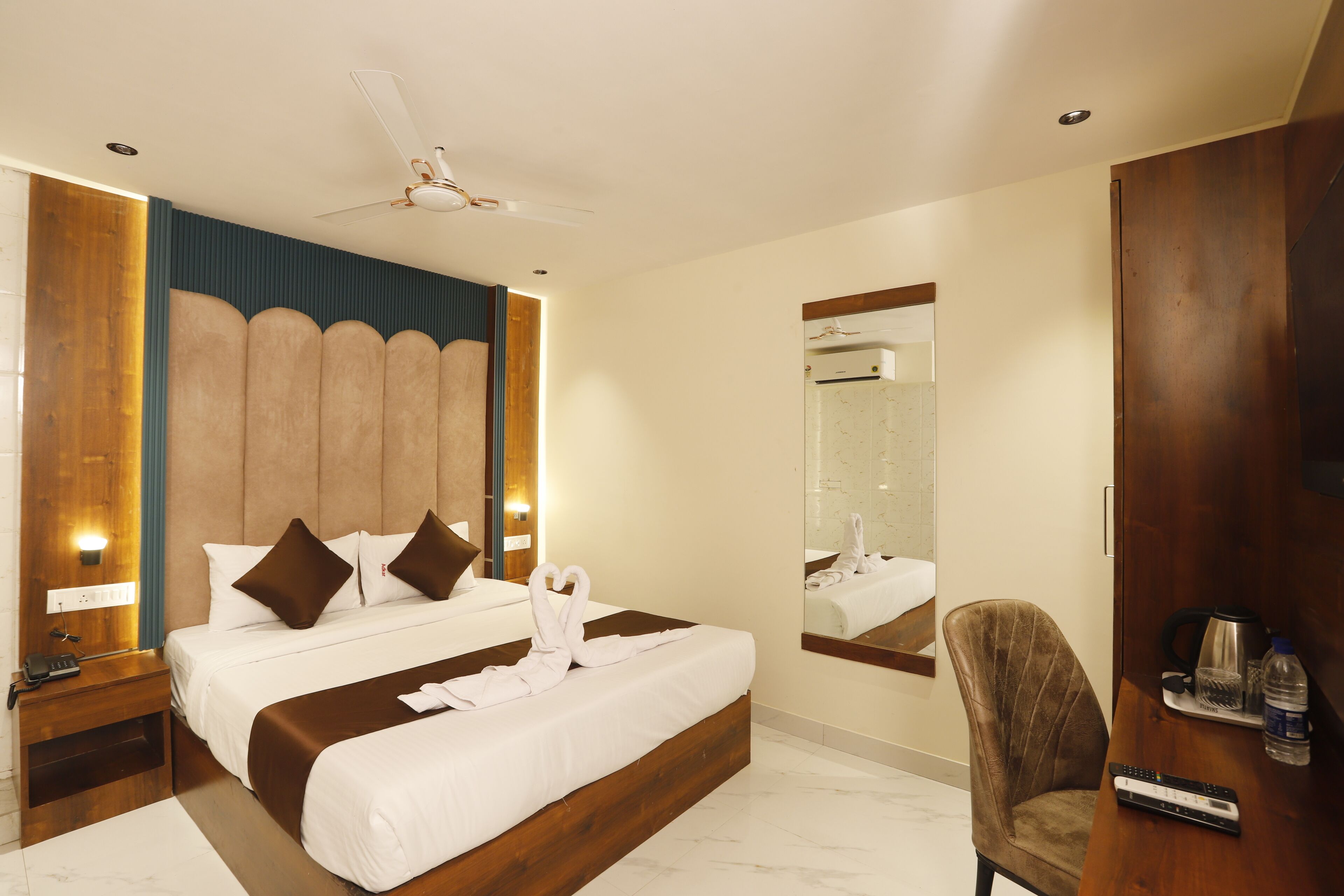 A hotel room with a large bed, a wooden headboard, a ceiling fan, a bedside table, a chair, and a mirror.