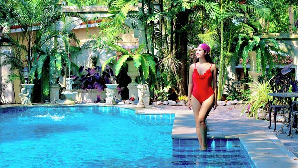 Una mujer con traje de baño rojo de pie junto a una piscina con fuentes decorativas y exuberante vegetación tropical.