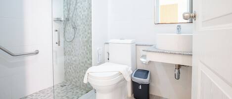 A modern bathroom with a glass shower enclosure, a white toilet, a sink with a mirror, and a trash bin.
