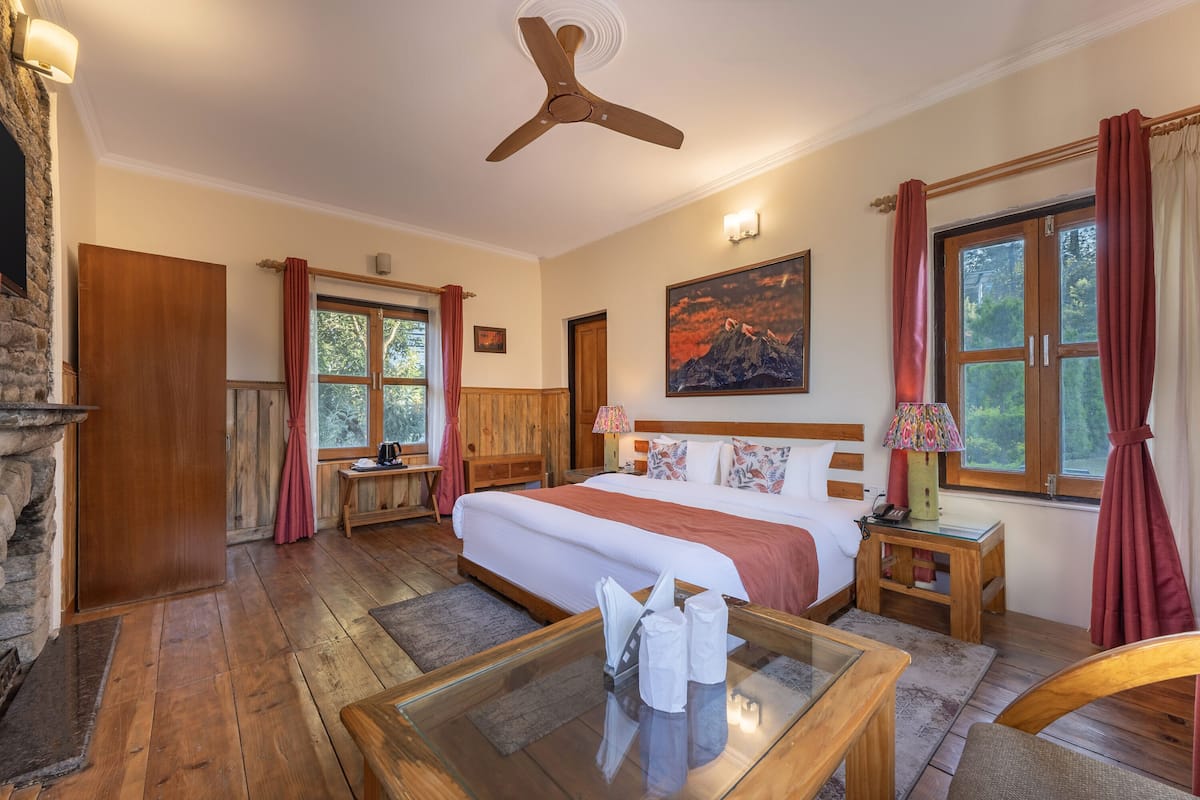 A hotel room with a large bed, wooden flooring, a stone fireplace, a ceiling fan, and a painting on the wall.