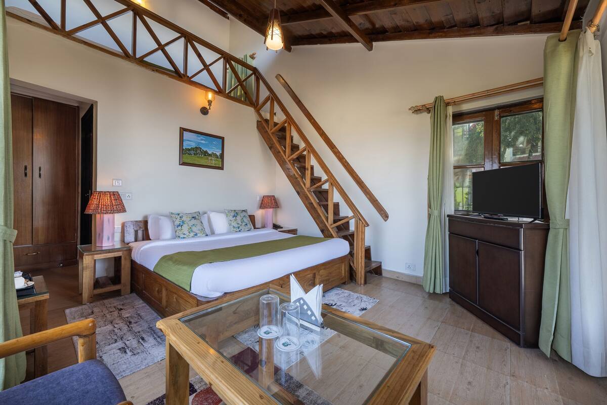 A bedroom with a bed, wooden staircase, a television, and a glass table.