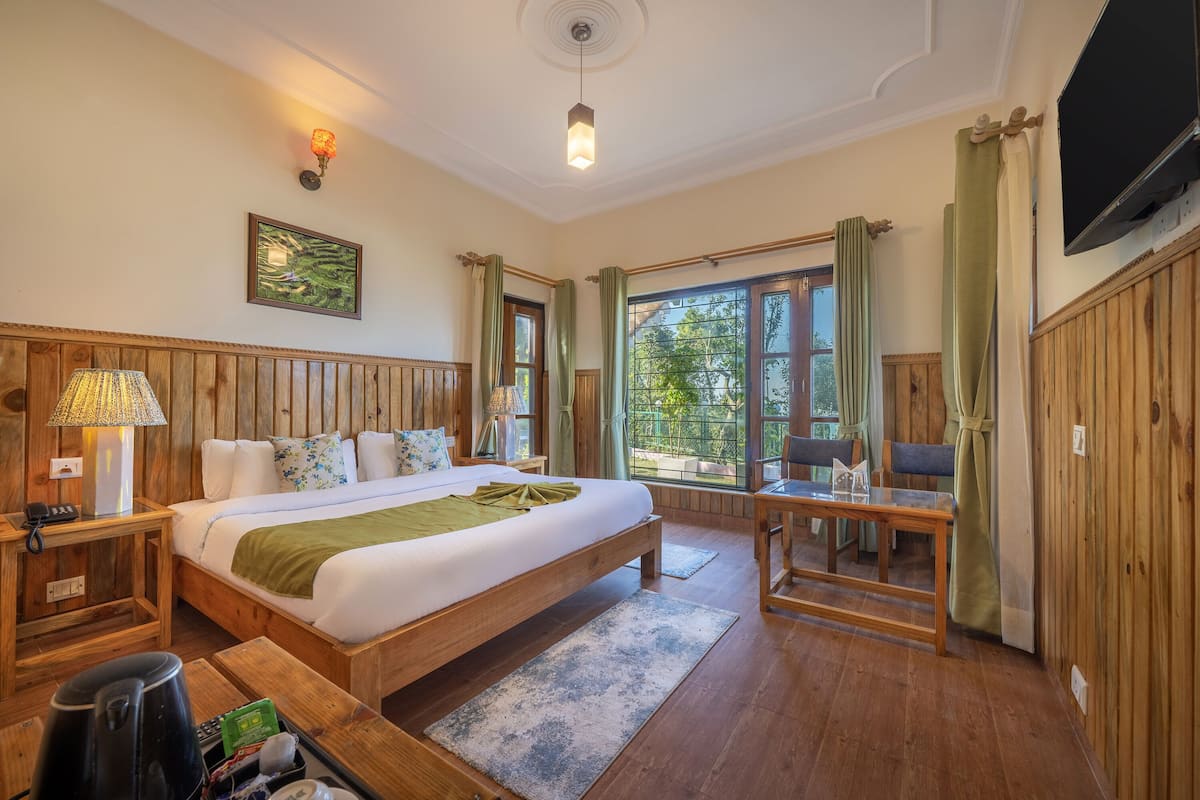 A wooden-furnished bedroom with a bed, bedside table, lamp, and a view of the outdoors.